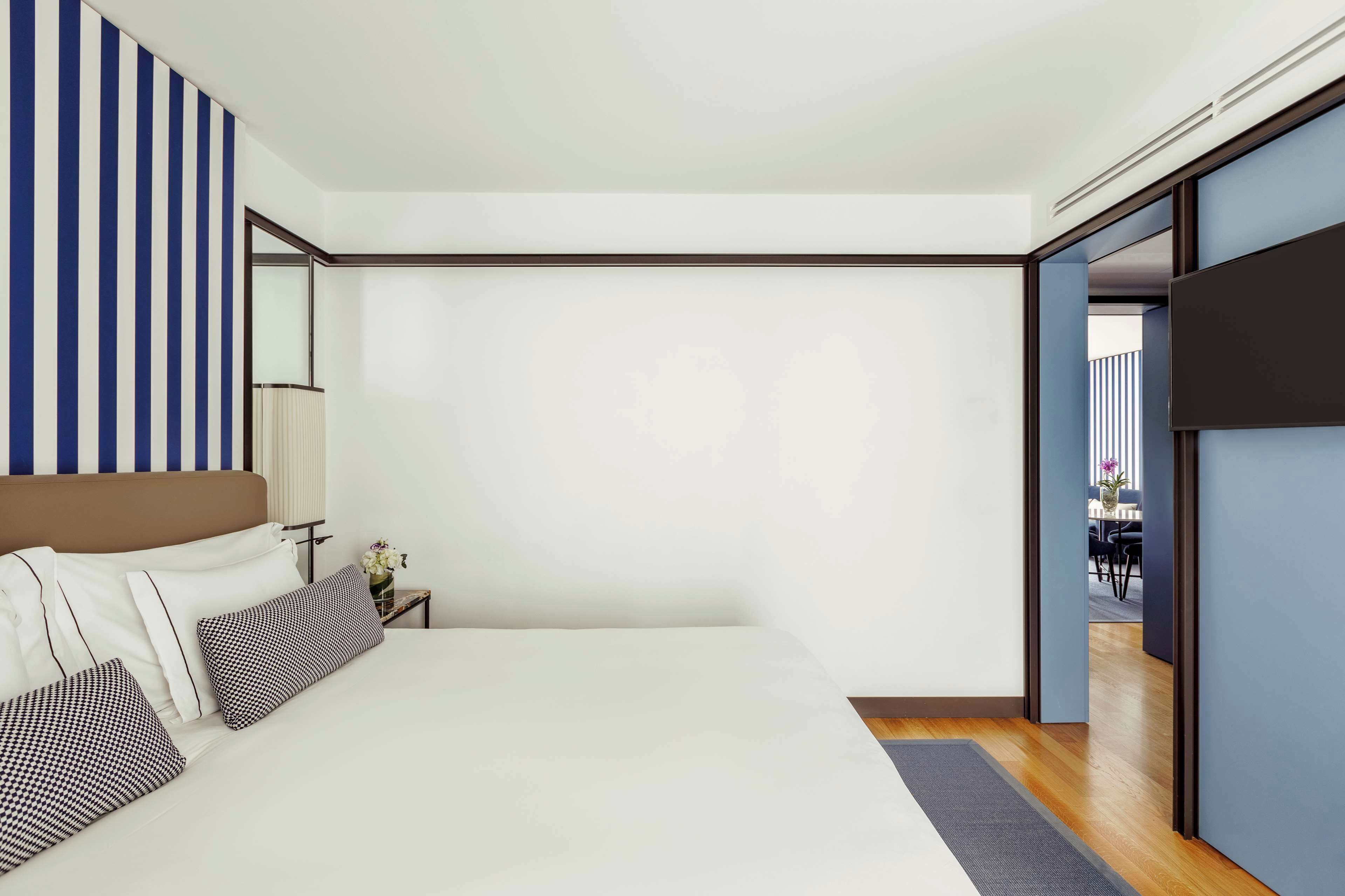 A modern bedroom with a large bed, a bedside table with a lamp, and a view into a dining area.