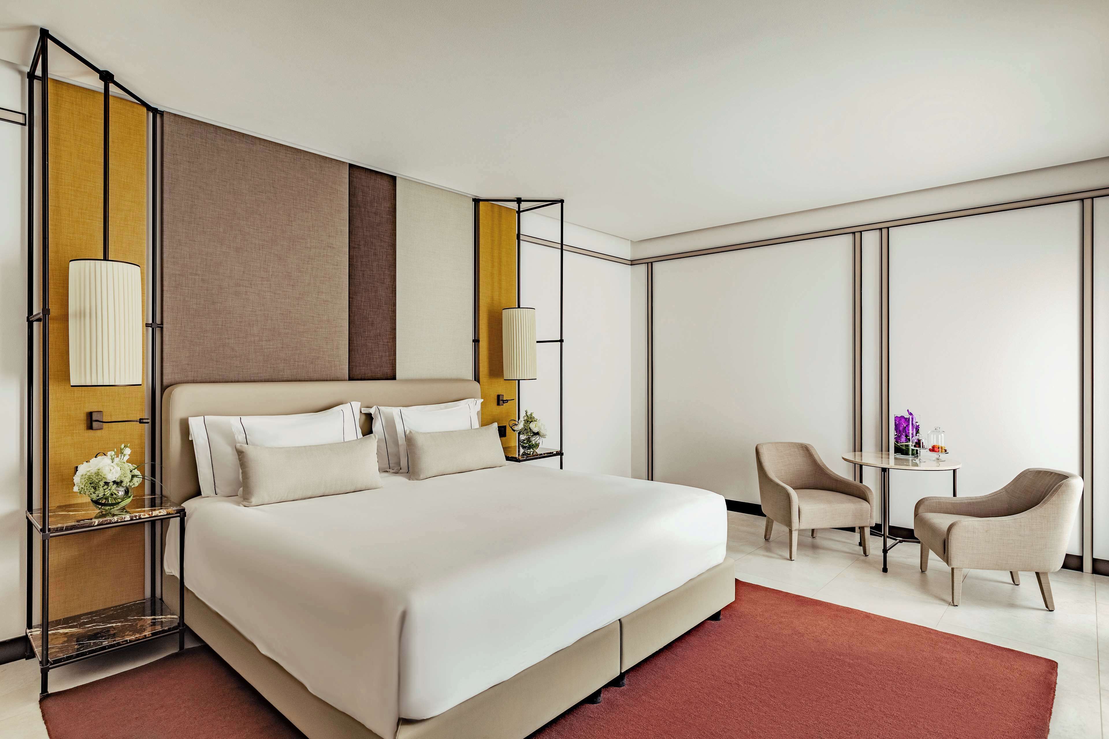 Una habitación de hotel moderna con una cama grande, dos mesitas de noche con lámparas, una pequeña mesa redonda con sillas y un amplio ventanal.