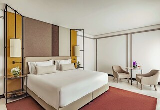 A modern hotel room with a large bed, two bedside tables with lamps, a small round table with chairs, and a large window.