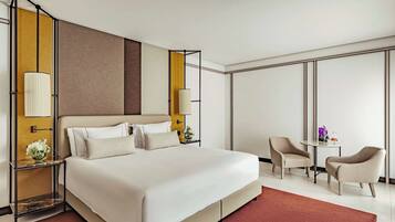 Una habitación de hotel moderna con una cama grande, dos mesitas de noche con lámparas, una pequeña mesa redonda con sillas y un amplio ventanal.