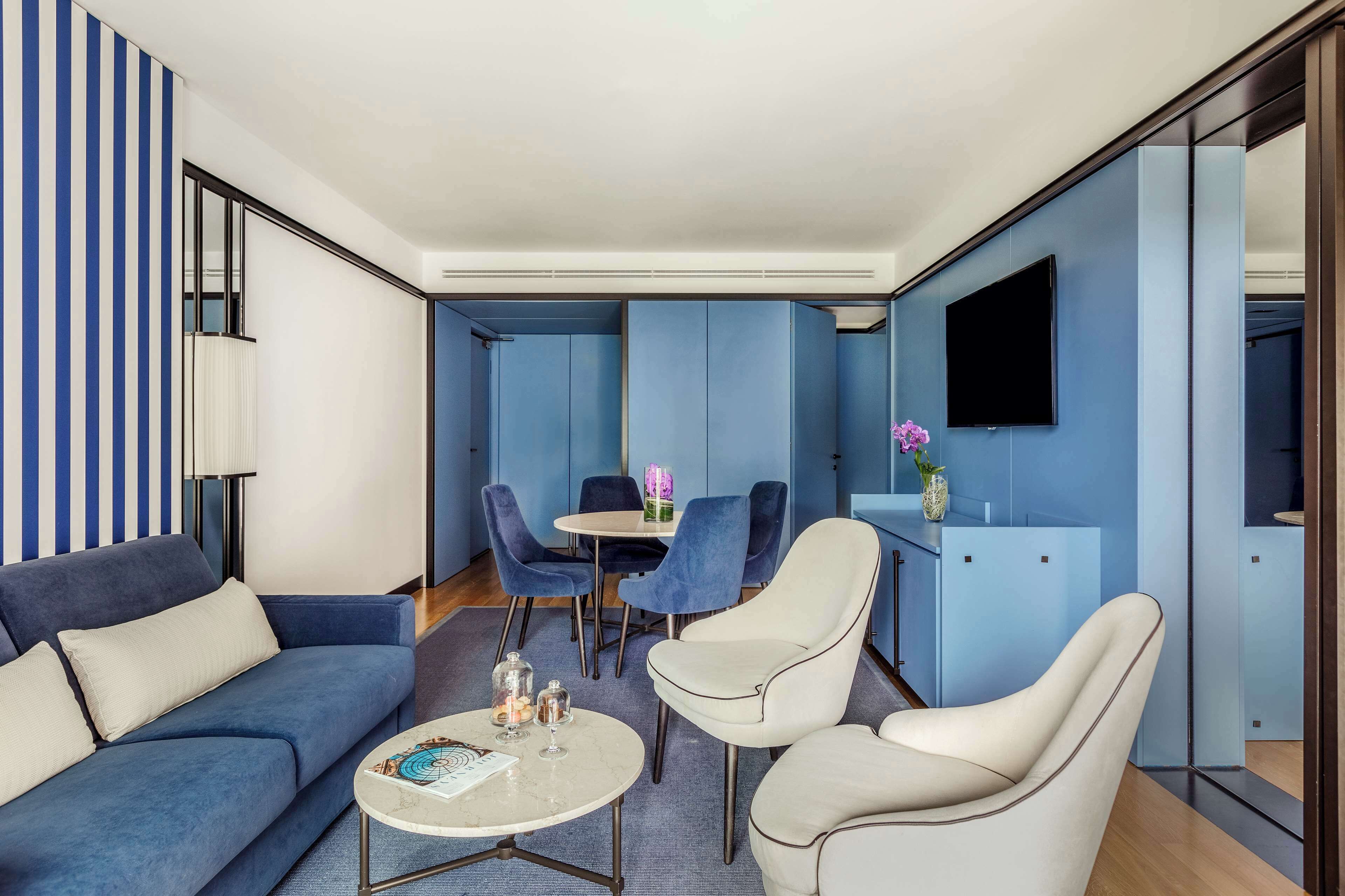 Una habitación de hotel moderna con una pared a rayas azules, un sofá, una mesa de comedor con sillas y un televisor de pantalla plana montado en la pared.