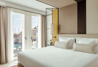 A hotel room with a large bed, a bedside table with a lamp, a view of the outside, and a balcony with a pool.