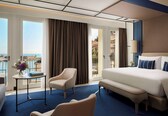 A hotel room with a large bed, a chair, a small table, and a view of the sea.