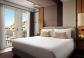 A hotel room with a large bed, a balcony with a view, a standing lamp, and a nightstand with a vase of flowers.