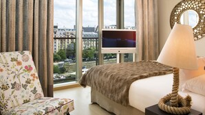 Hotelkamer met een groot raam, een bed, een televisie, een lamp en een bloemen fauteuil.