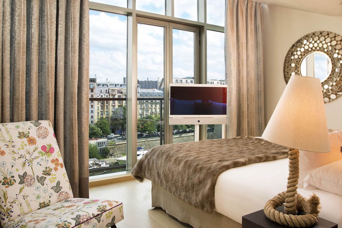 Habitación de hotel con un ventanal grande, una cama, un televisor, una lámpara y un sillón con estampado floral.