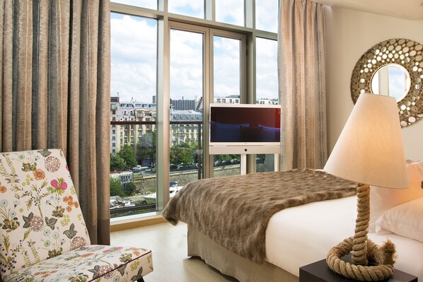 Habitación de hotel con un ventanal grande, una cama, un televisor, una lámpara y un sillón con estampado floral.