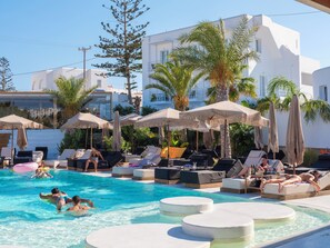 Zona de piscina con sillones de descanso y sombrillas, rodeada de palmeras y un edificio blanco.