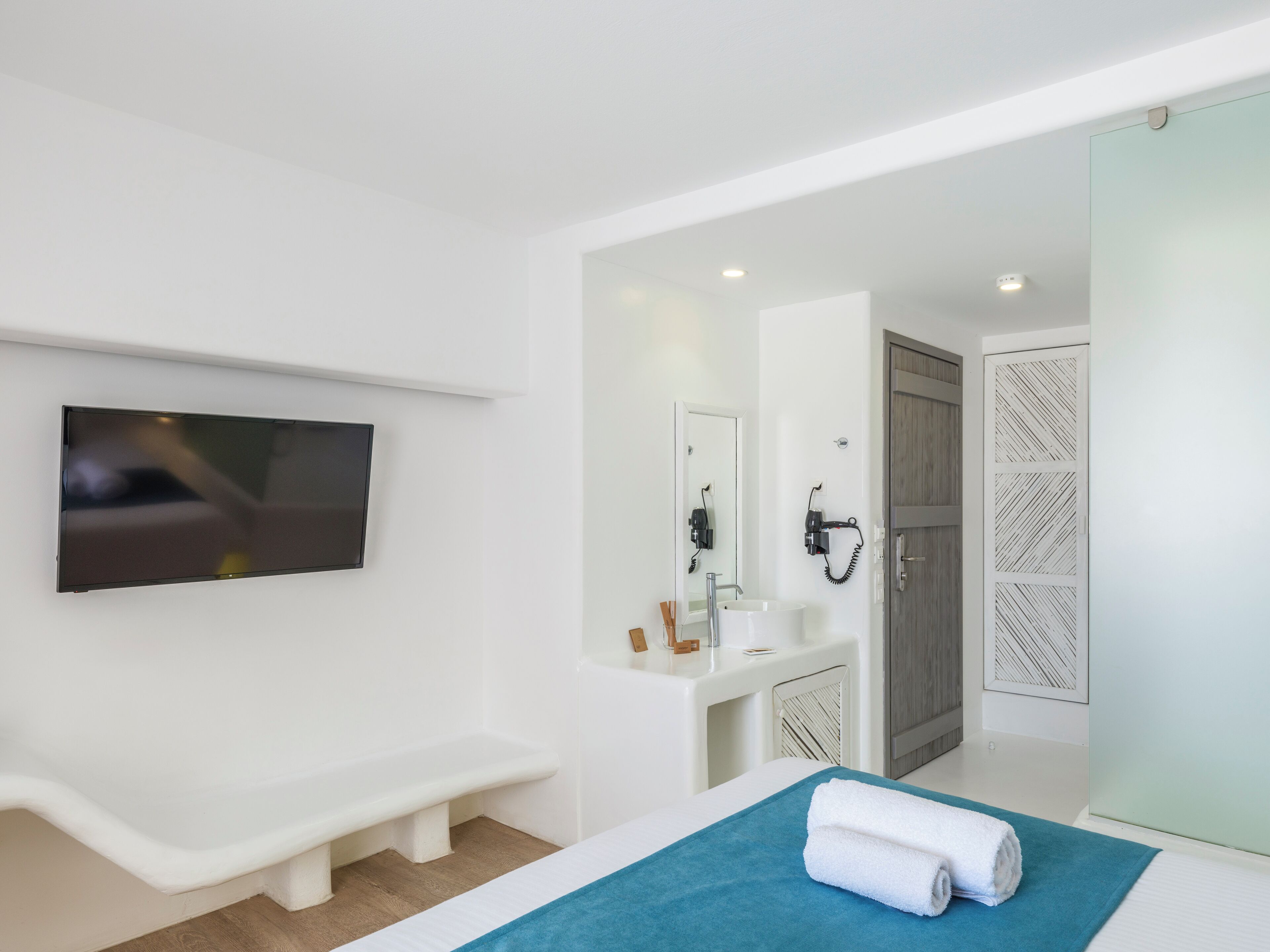 A modern hotel room with a flat-screen TV mounted on the wall, a white bench, a sink with a mirror, and a door with a patterned panel.