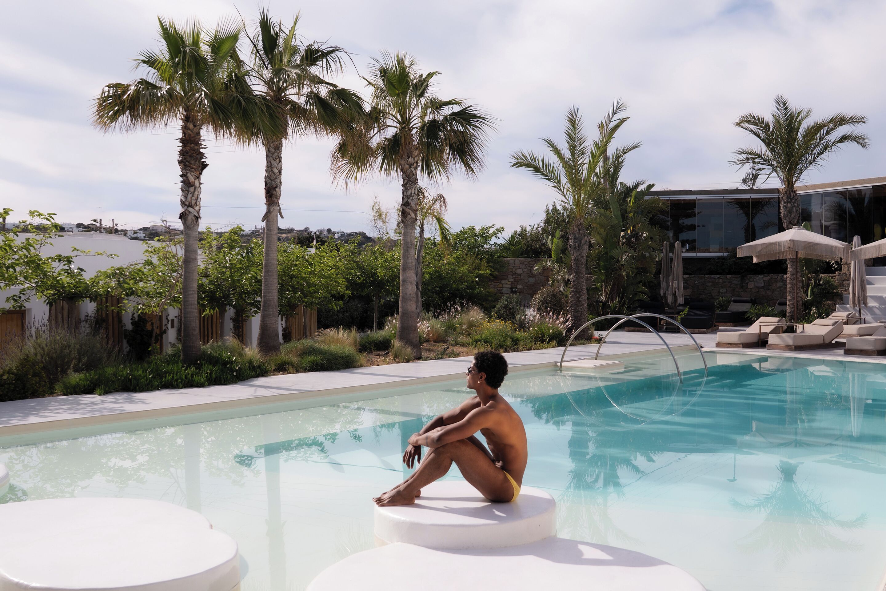 Un hombre sentado junto a una piscina, con palmeras y un edificio moderno al fondo.