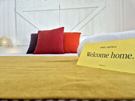 Una habitación de hotel con cama, almohadas, un cartel de bienvenida y una lámpara.