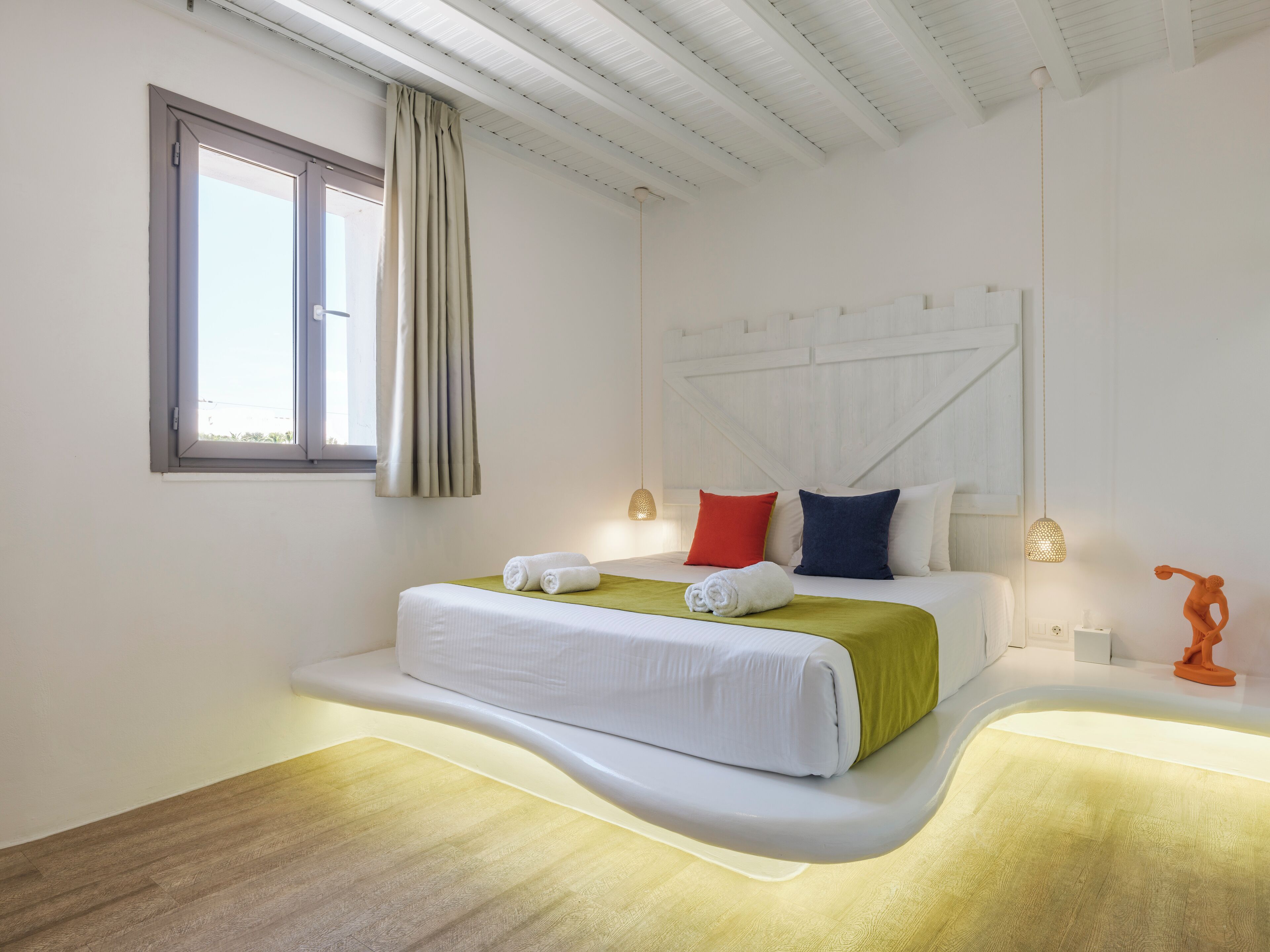 A modern bedroom with a curved bed, a window with curtains, and a unique orange sculpture.