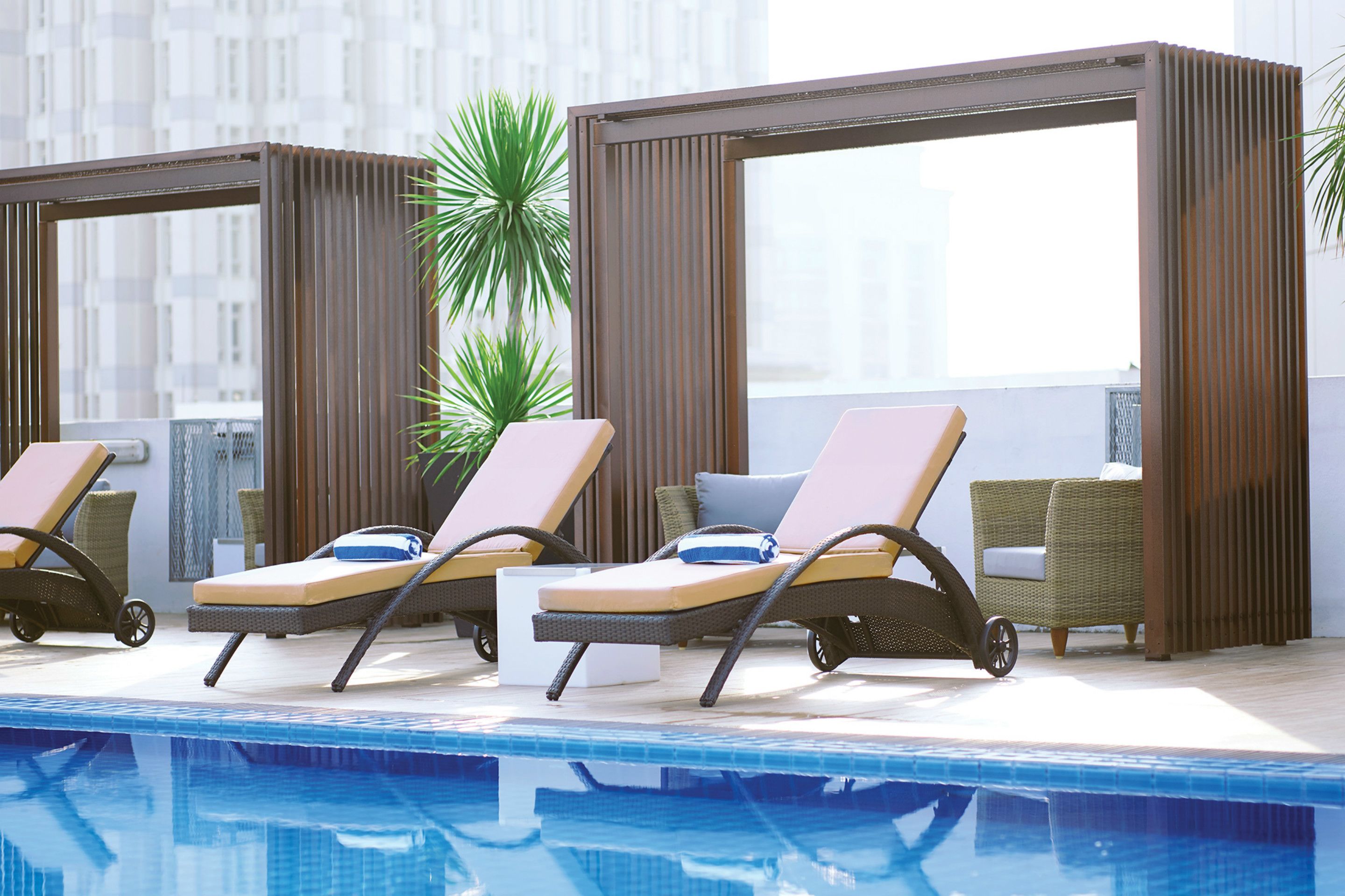 A rooftop pool area with lounge chairs and a wicker sofa, surrounded by wooden panels and a cityscape view.