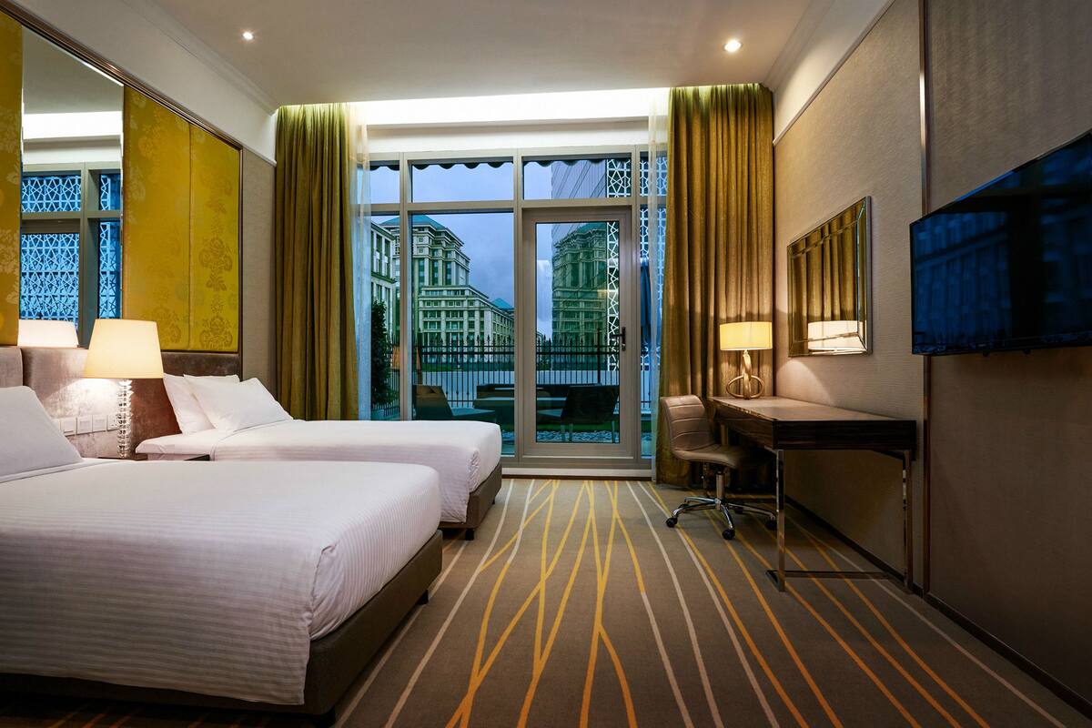 Een hotelkamer met twee bedden, een bureau, een stoel en een groot raam met uitzicht op gebouwen.