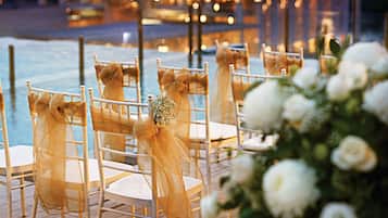 A wedding setup with chairs and a floral arrangement on a wooden deck by a body of water, with a dome-shaped structure in the background.