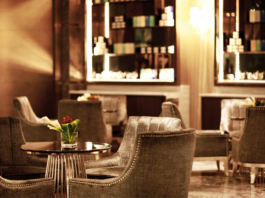 A seating area with upholstered chairs and a small table, a vase with flowers, and a bar with shelves of bottles.