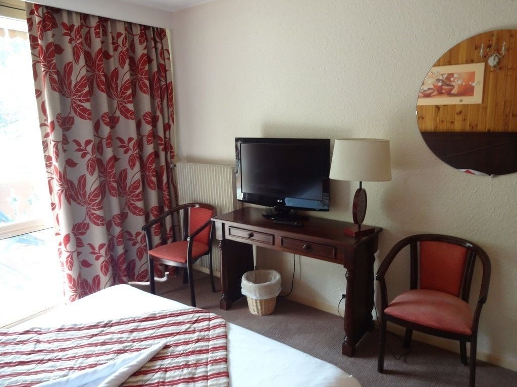 Habitación de hotel con cama, escritorio con televisor, dos sillones, lámpara y ventana con cortinas.