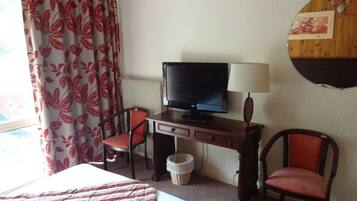 Habitación de hotel con cama, escritorio con televisor, dos sillones, lámpara y ventana con cortinas.