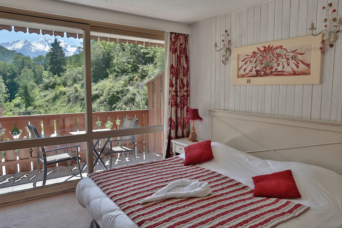 Habitación de hotel con balcón, cama con ropa de cama roja y blanca, un cuadro en la pared y vistas a la montaña.