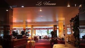 Vista del interior del restaurante Le Verseau, con un ambiente cálido, que muestra una zona central de comedor con mesas y sillas, una zona de bar con mostrador, y elementos decorativos como plantas y obras de arte.