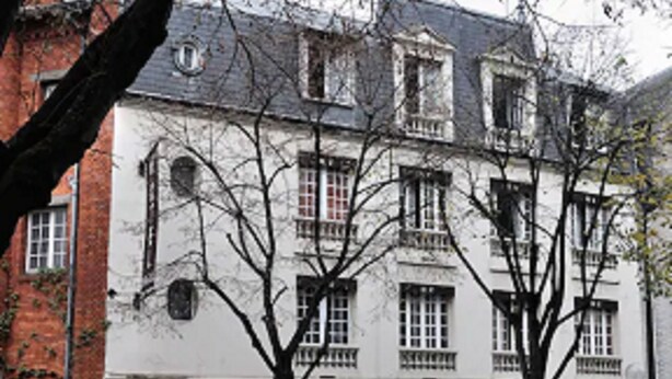 Un bâtiment doté de nombreuses fenêtres et d’une porte, entouré d’arbres et d’un mur de briques.