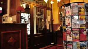 Un hall d’accueil d’hôtel avec un comptoir de réception en bois, une moquette rouge et un mur recouvert de brochures de voyage.