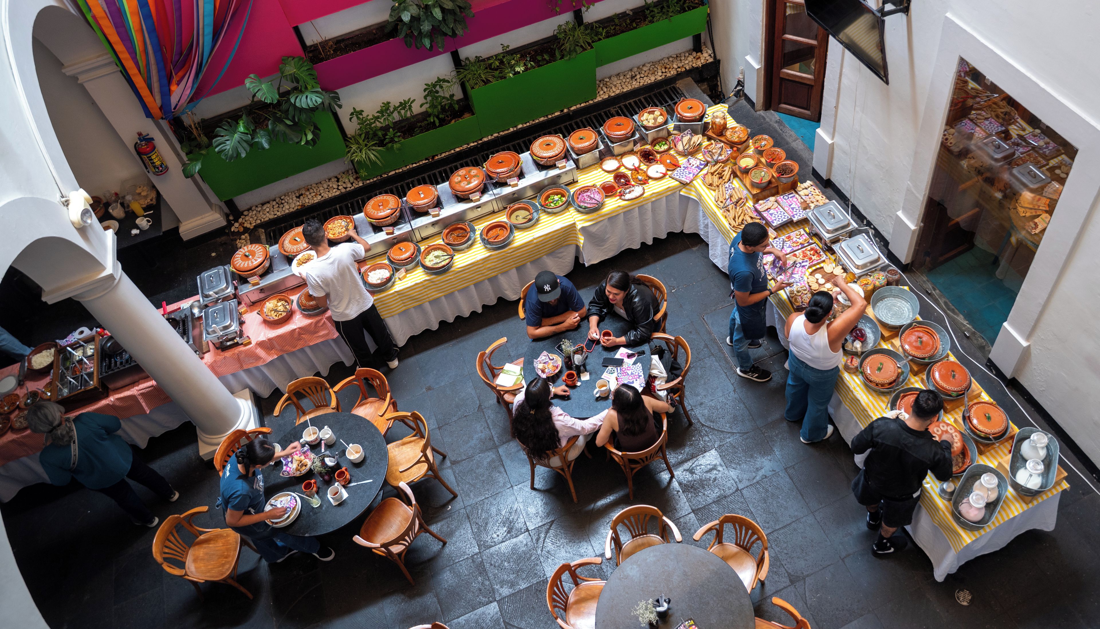 Un comedor con varias mesas y sillas, una barra de buffet con diversos alimentos, y una pared verde con plantas.