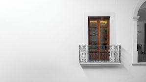 A white building with a window featuring a wooden door and a balcony with a wrought iron railing. - Del Carmen Concept Hotel (Guadalajara)
