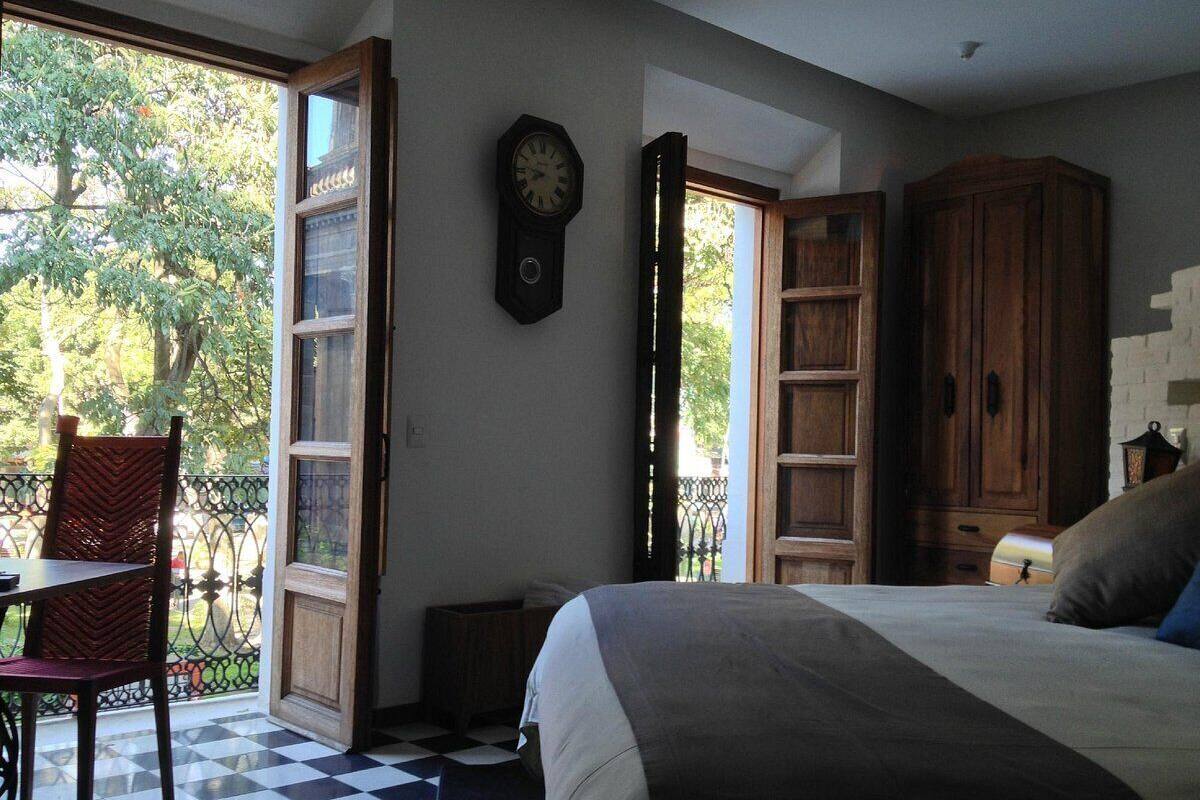 A bedroom with a bed, a wooden wardrobe, a clock on the wall, and a window with a view of trees.