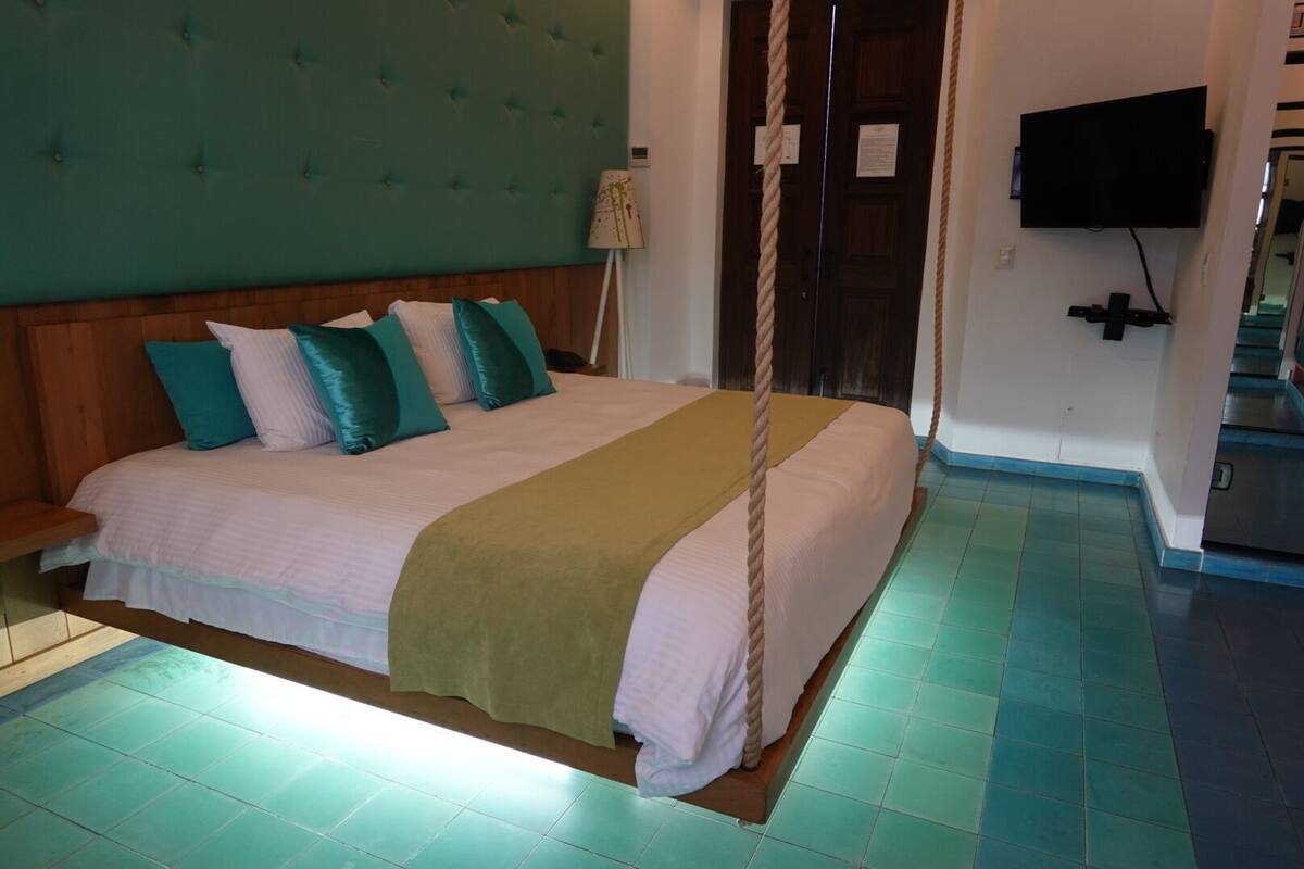 A bed with a headboard, multiple pillows, a bedside lamp, a television mounted on the wall, and a staircase in the background.