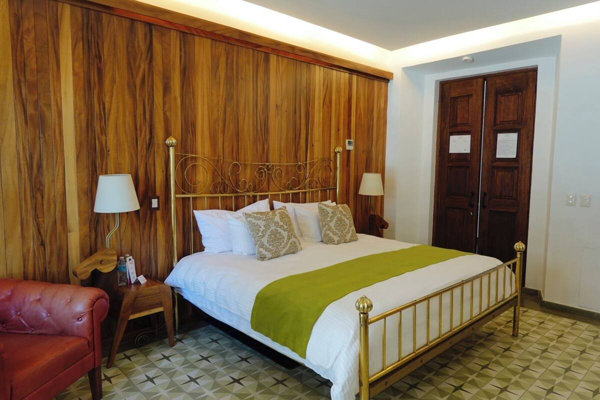 A hotel room with a wooden panel wall, a bed with a green blanket, two bedside lamps, and a red leather chair.