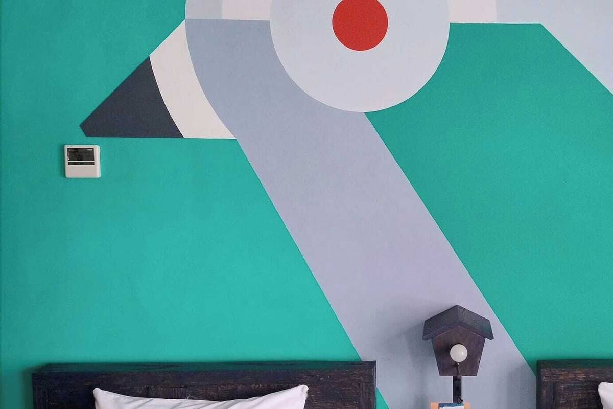 A room with a geometric wall mural, a bed with white bedding, and a small bedside table with a lamp.