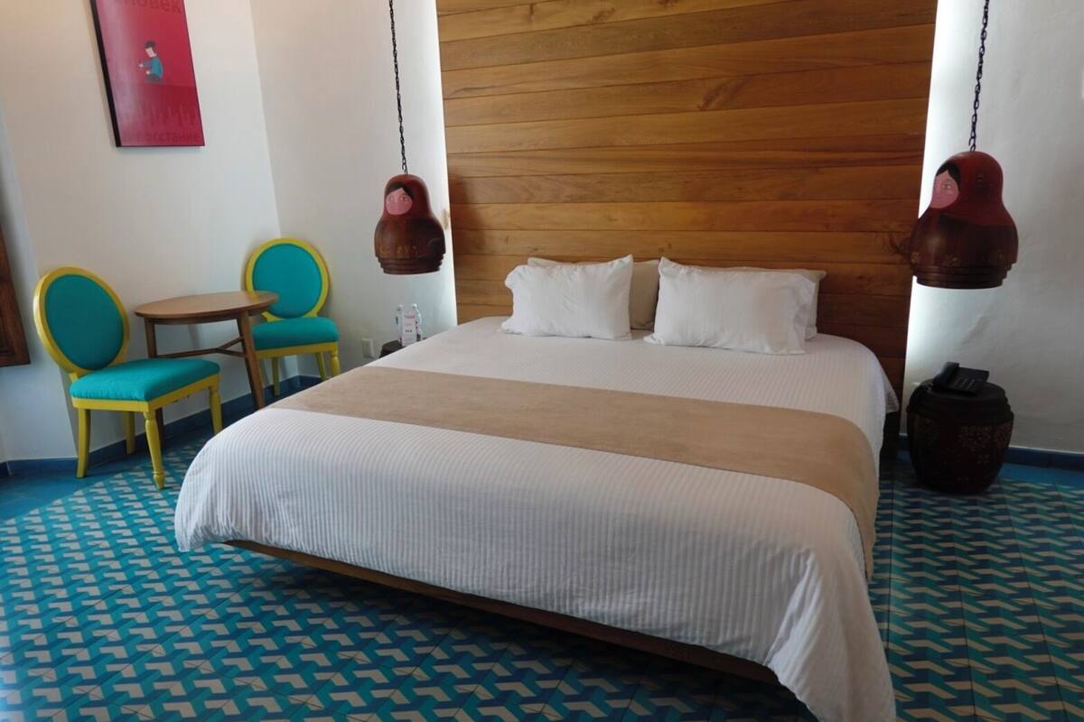 A hotel room with a large bed, a wooden headboard, two hanging lamps, a small table, and two chairs.