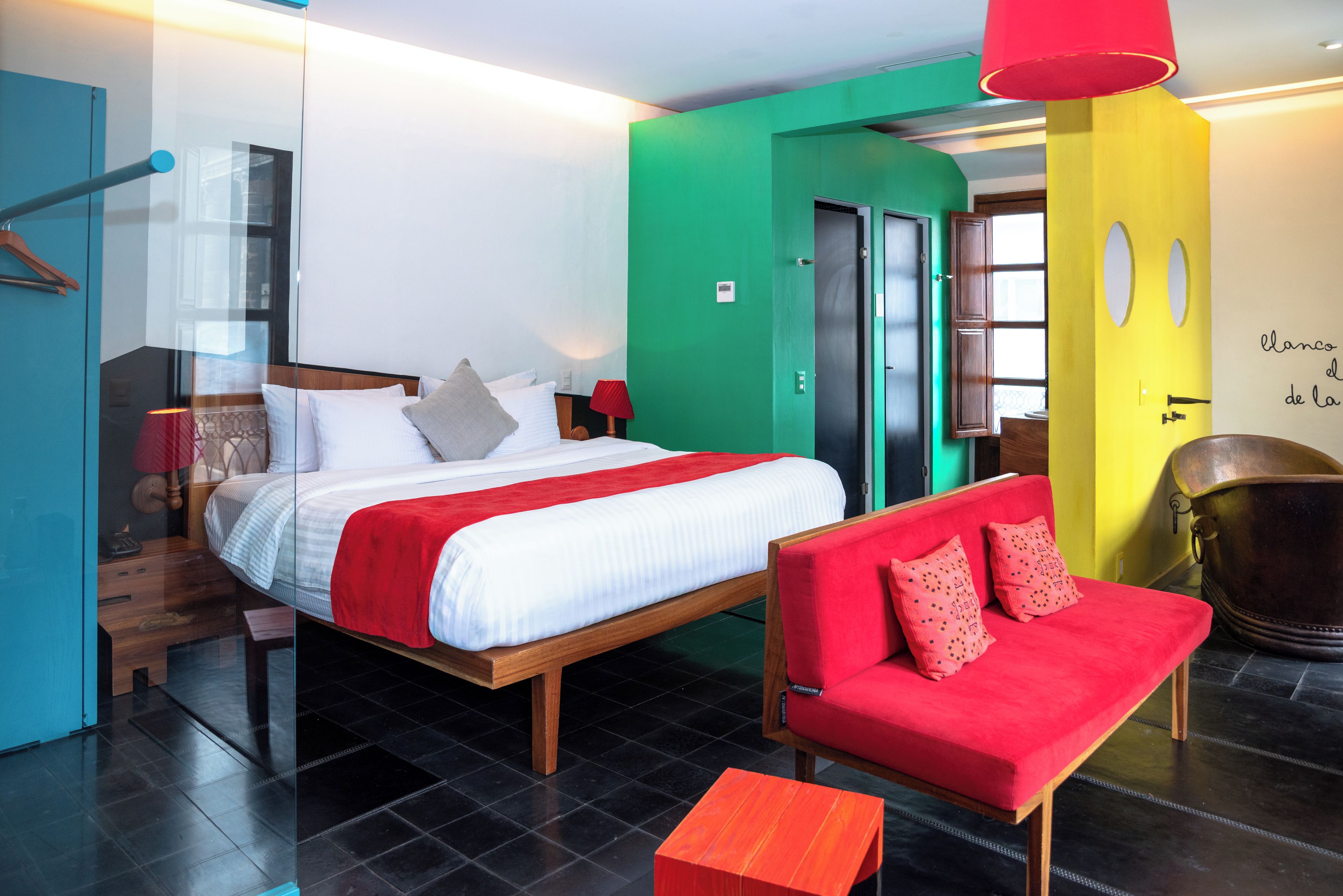 Habitación de hotel con cama, sofá rojo, mesa de madera y baño con pared amarilla.