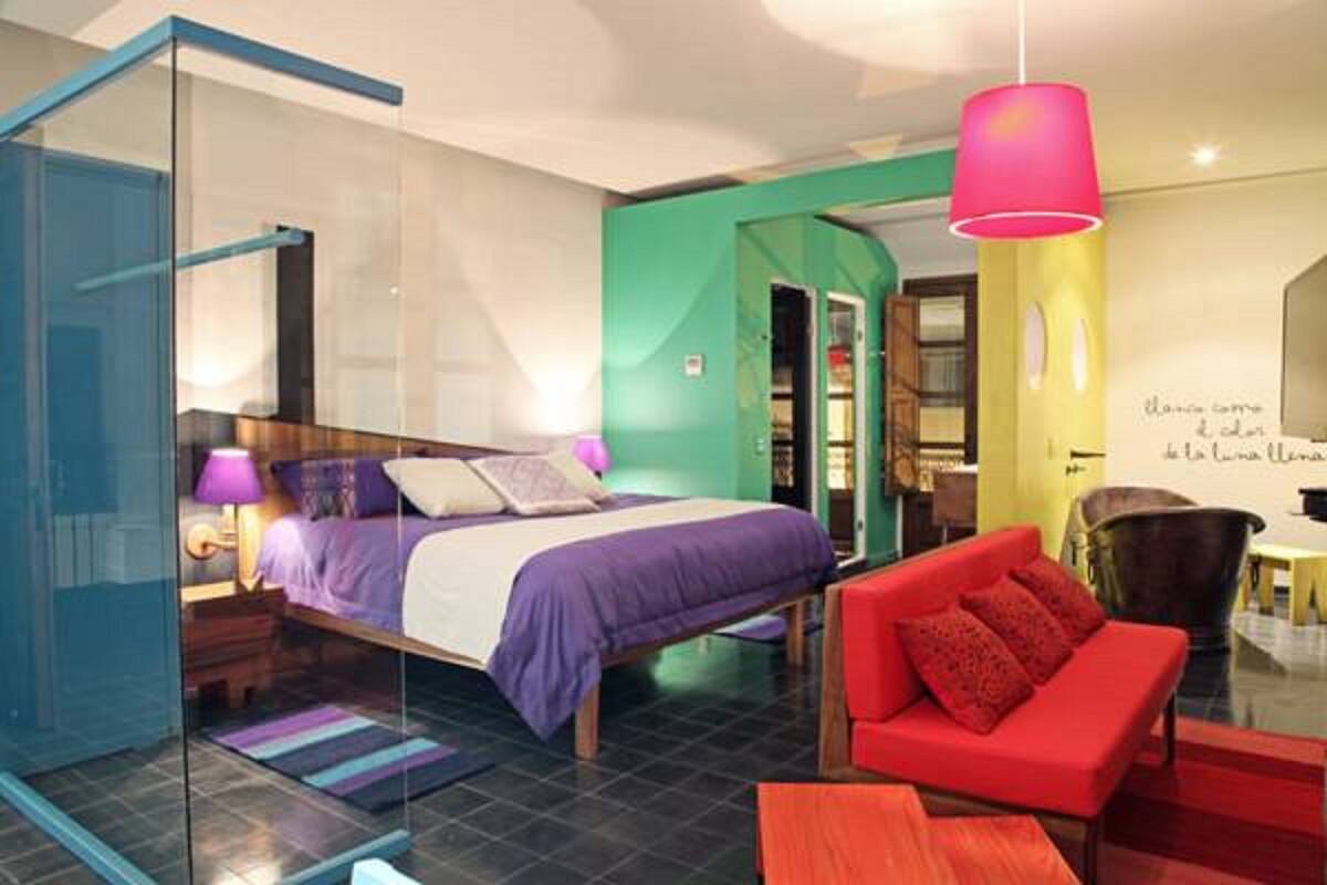 A modern hotel room with a glass partition, a bed with purple bedding, a red sofa, and a pink pendant light.