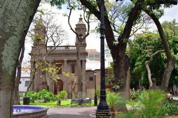 Un edificio histórico con una torre central, rodeado de árboles y una fuente.