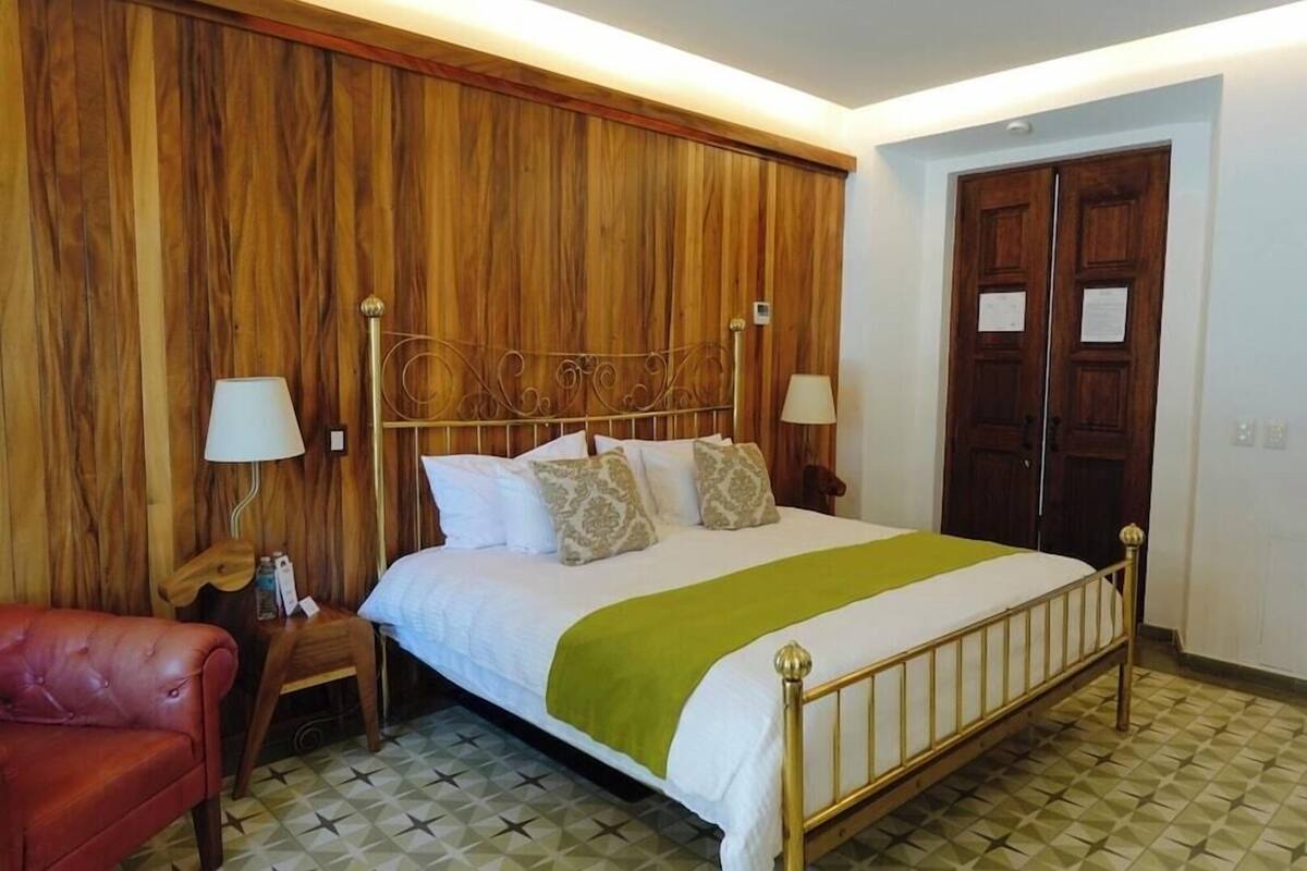A bedroom with a wooden headboard, a bed with white bedding and a green throw, a red leather chair, and a wooden nightstand with a lamp.