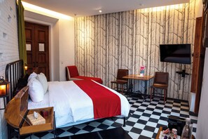 Hotelkamer met een geruit vloerkleed, een bed met wit en rode beddengoed, een bureau met stoel, een televisie aan de muur en een houten deur.