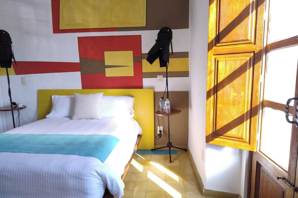 A hotel room with a bed, a small table with water bottles, a lamp, and a door with a wooden frame.