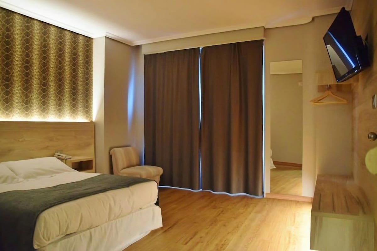 A hotel room with a bed, a chair, a TV mounted on the wall, and a large window with curtains.