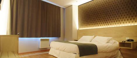 A modern hotel room with a large bed, a wooden headboard, a bedside table, and a large window with curtains.