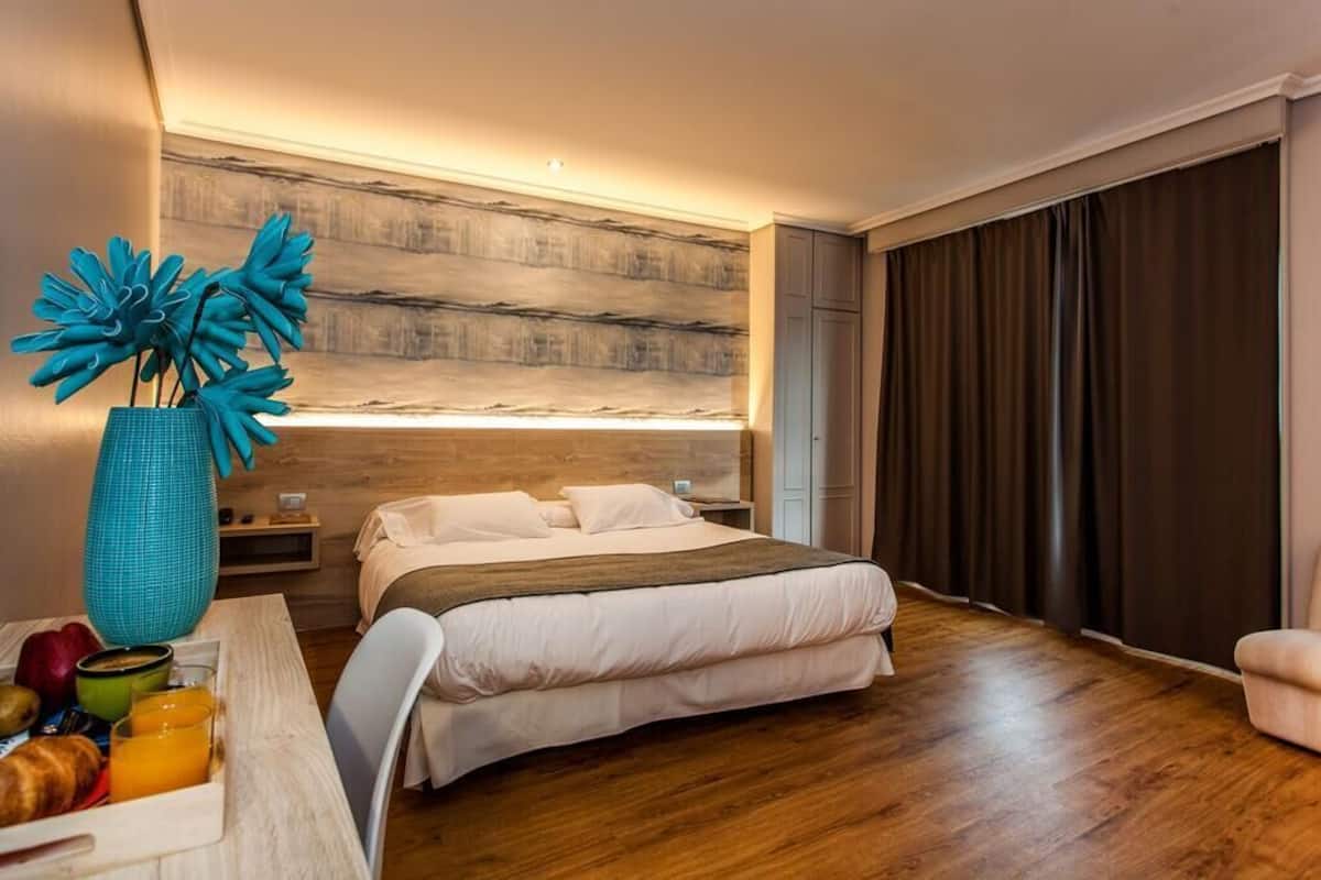 Un dormitorio moderno con cabecera de madera, cama con ropa de cama blanca, una mesa de comedor con alimentos para el desayuno y un jarrón grande con flores azules.