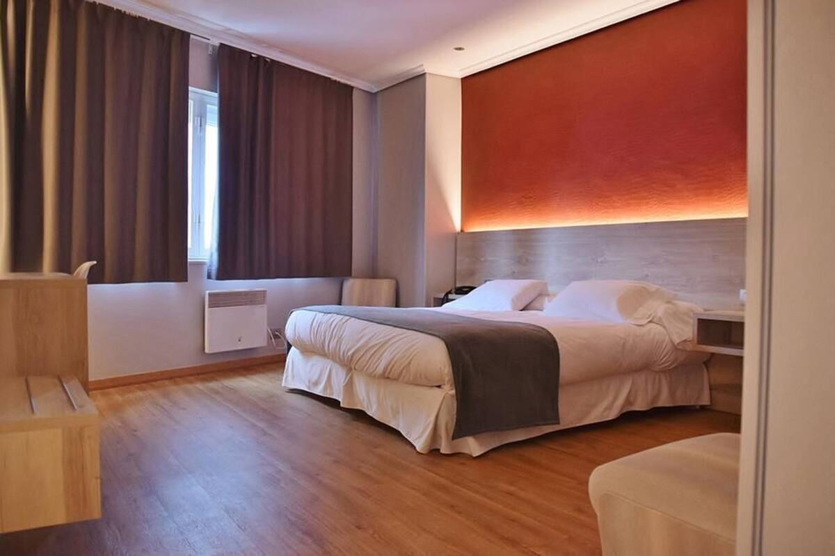 Una habitación de hotel moderna con una cama grande, una mesita de noche, una silla y una ventana con cortinas.