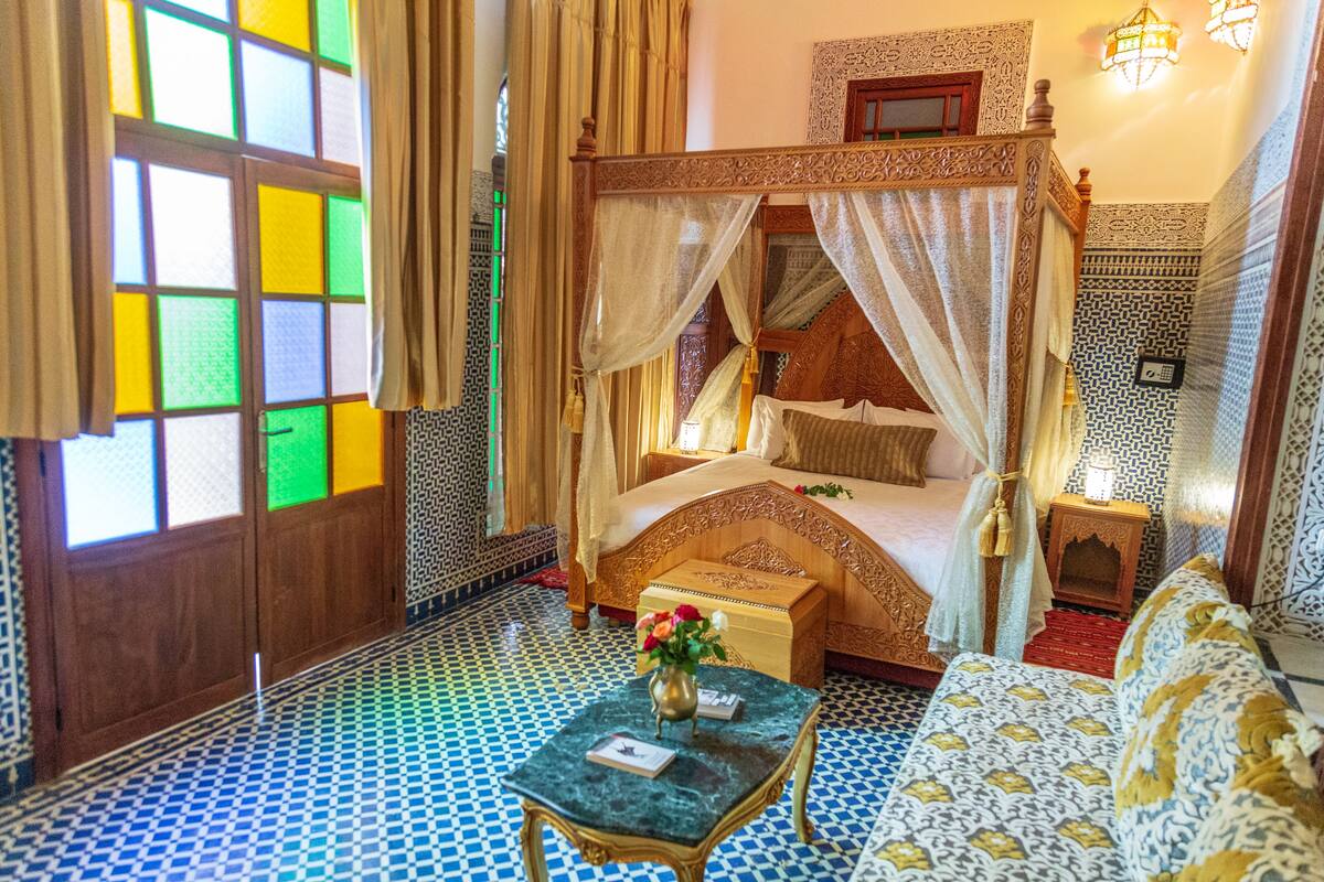 A four-poster bed with a canopy, a small table with a vase, and a patterned sofa in a room with blue and white tiled floors and a large window with colorful panes.