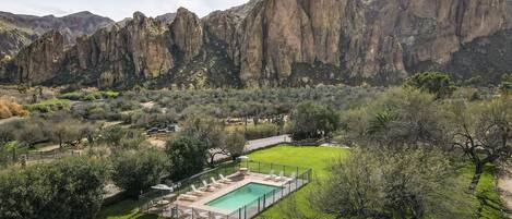 A pool area with lounge chairs and a fenced deck, surrounded by lush greenery and mountains in the background.