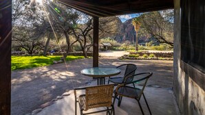 Un porche techado con una mesa de vidrio y sillas de mimbre, con vista a un jardín ajardinado y montañas al fondo.