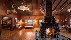 Interior rústico de una cabaña con chimenea de piedra, pisos de madera, mobiliario de cuero y trofeos de animales en las paredes.