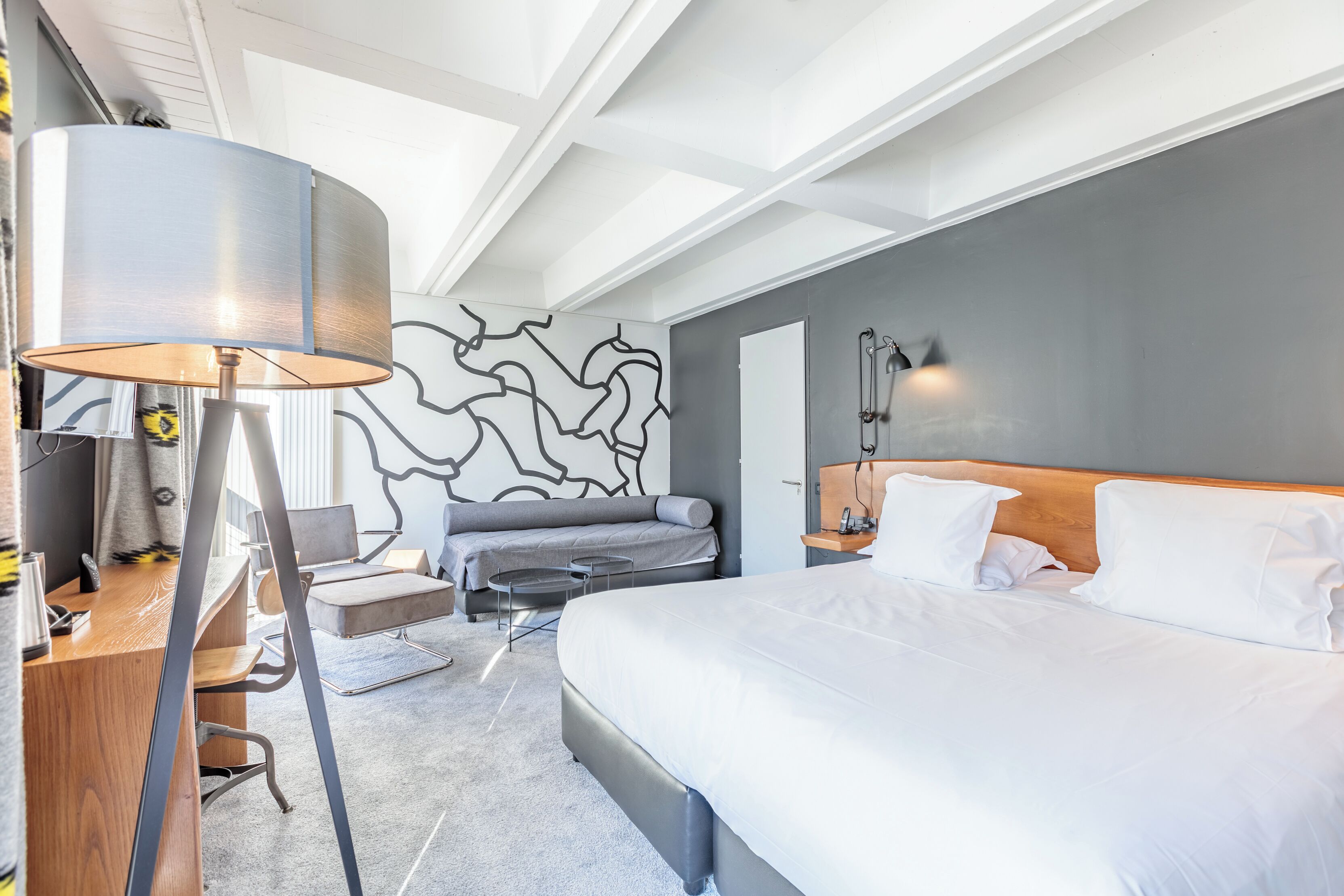 Une chambre d’hôtel moderne, équipée d’un lit, d’un bureau avec une lampe, d’un canapé et d’un mur décoré d’œuvres d’art abstraites.