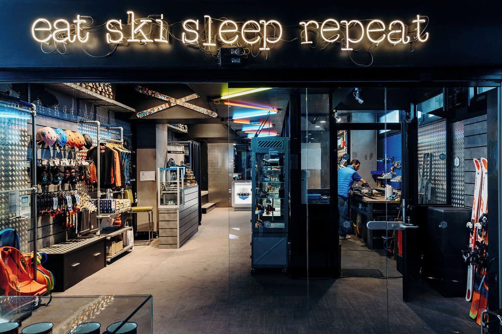 A ski shop interior with a neon sign reading "eat ski sleep repeat," shelves with ski gear, and a person working at a counter.