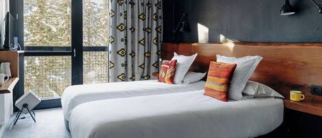 A hotel room with a large bed, a wooden headboard, a desk with a chair, a window with curtains, and a lamp on the wall.